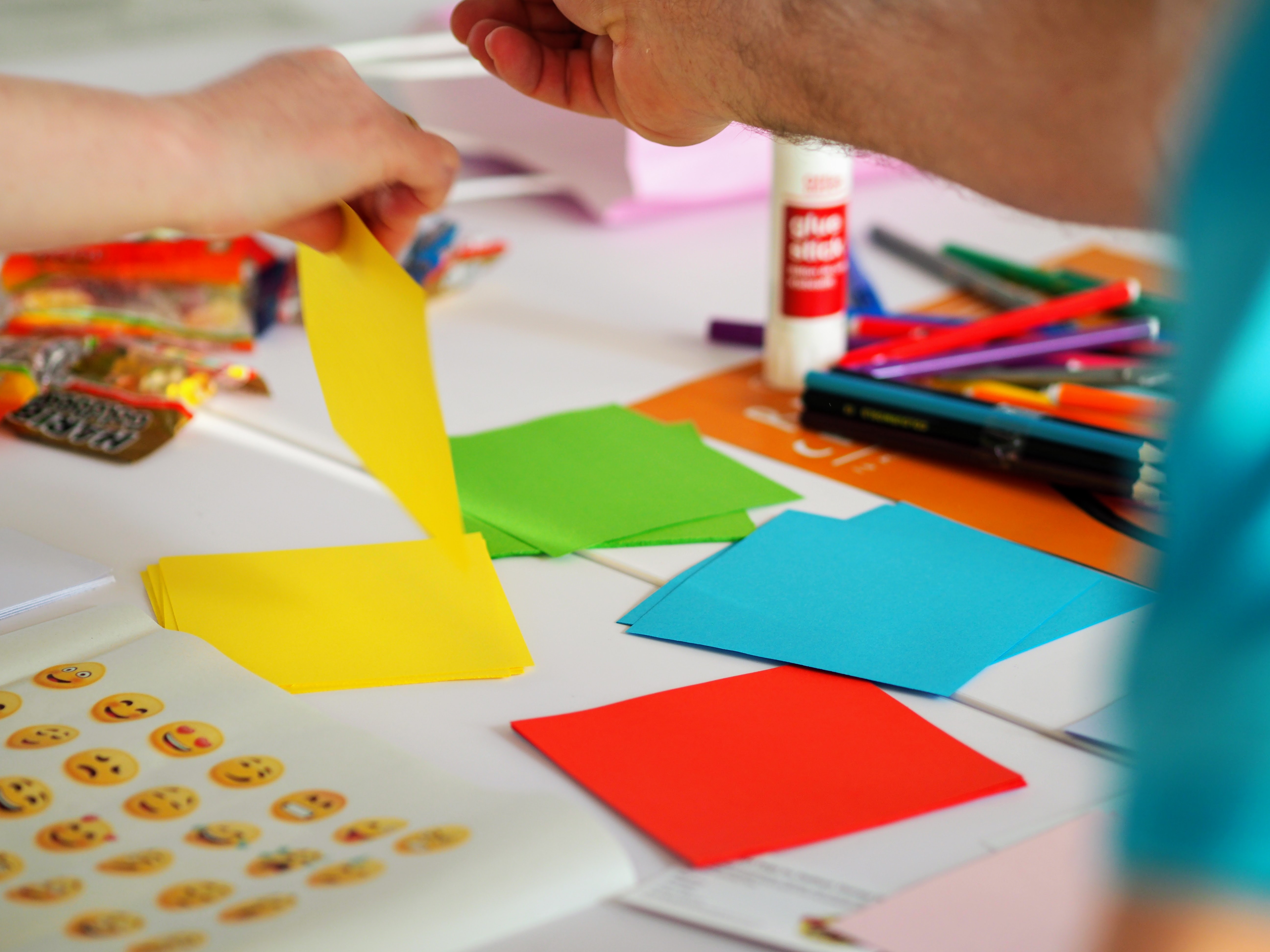 Colourful paper squares next to a pile of felt tip pens and a glue stick.